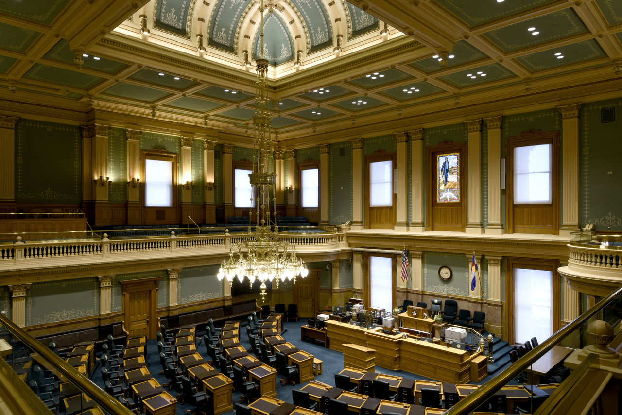 Modern view from the balcony showing the room surrounded by windows on two levels, a chandelier hanging from the skylight, and desks arranged in a half-circle around the main podium